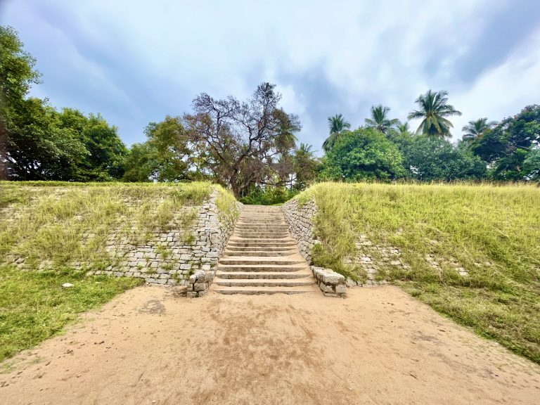 Stone staircase leading to Underground Shiva Temple, Hampi, Karnataka with tropical trees and a cloudy sky in the background.