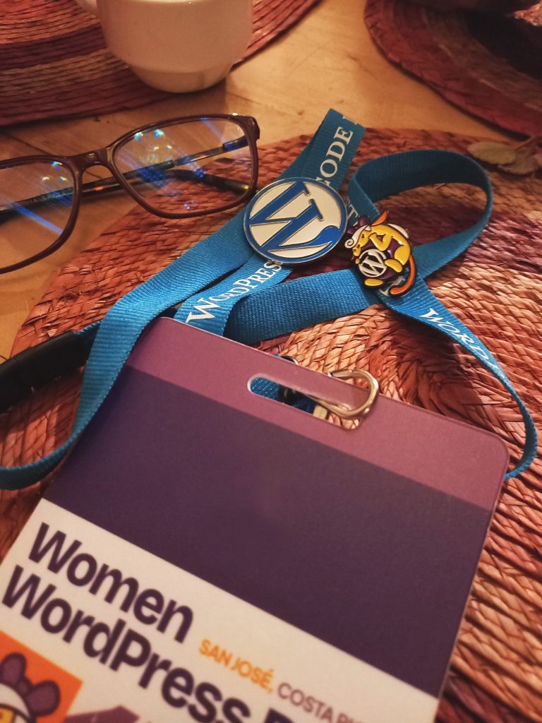 A pair of eyeglasses, a blue lanyard with WordPress branding, a conference badge reading “Women in WordPress,” a WordPress logo pin, and a wapuu pin, all placed on a woven table mat.