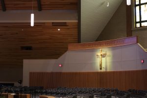 Inside a modern christian church, rows of chairs face a stage with a lit cross under "WORTHY IS THE LAMB" text.