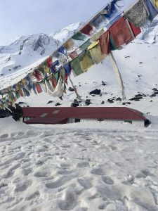 A view of Annapurna Base Camp reveals a snow-covered landscape adorned with colorful prayer flags above a red-roofed building against a backdrop of mountain peaks.