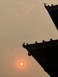 A hazy sunset with the orange sun visible through a misty sky, framed by the silhouettes of traditional Bagbhairav Temple in Kirtipur, Nepal