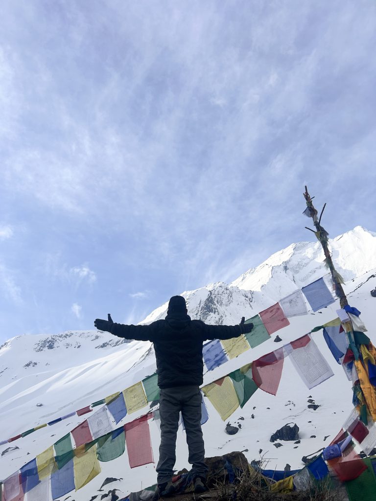 A person with arms outstretched facing a snow-covered mountain, with colorful prayer flags in the foreground against a cloudy blue sky in Annapurna Base Camp.