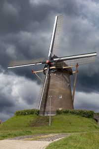 A traditional Dutch windmill in Wijchen, The Netherlands set against a backdrop of a dramatic cloudy sky. The windmill is atop a grassy hill with a cobblestone path leading up to it.