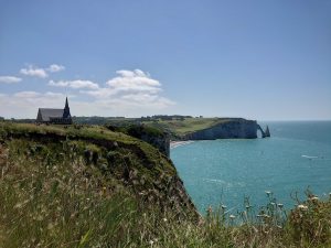 The stunning coastline of Étretat, France features a cliffside church, towering cliffs in the background, and a beautiful blue sky.
