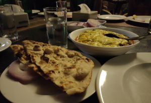 A dinner setting with a plate of naan bread, punjabi dish with cheese topping, glasses of water, and additional tableware on a dark restaurant table.
