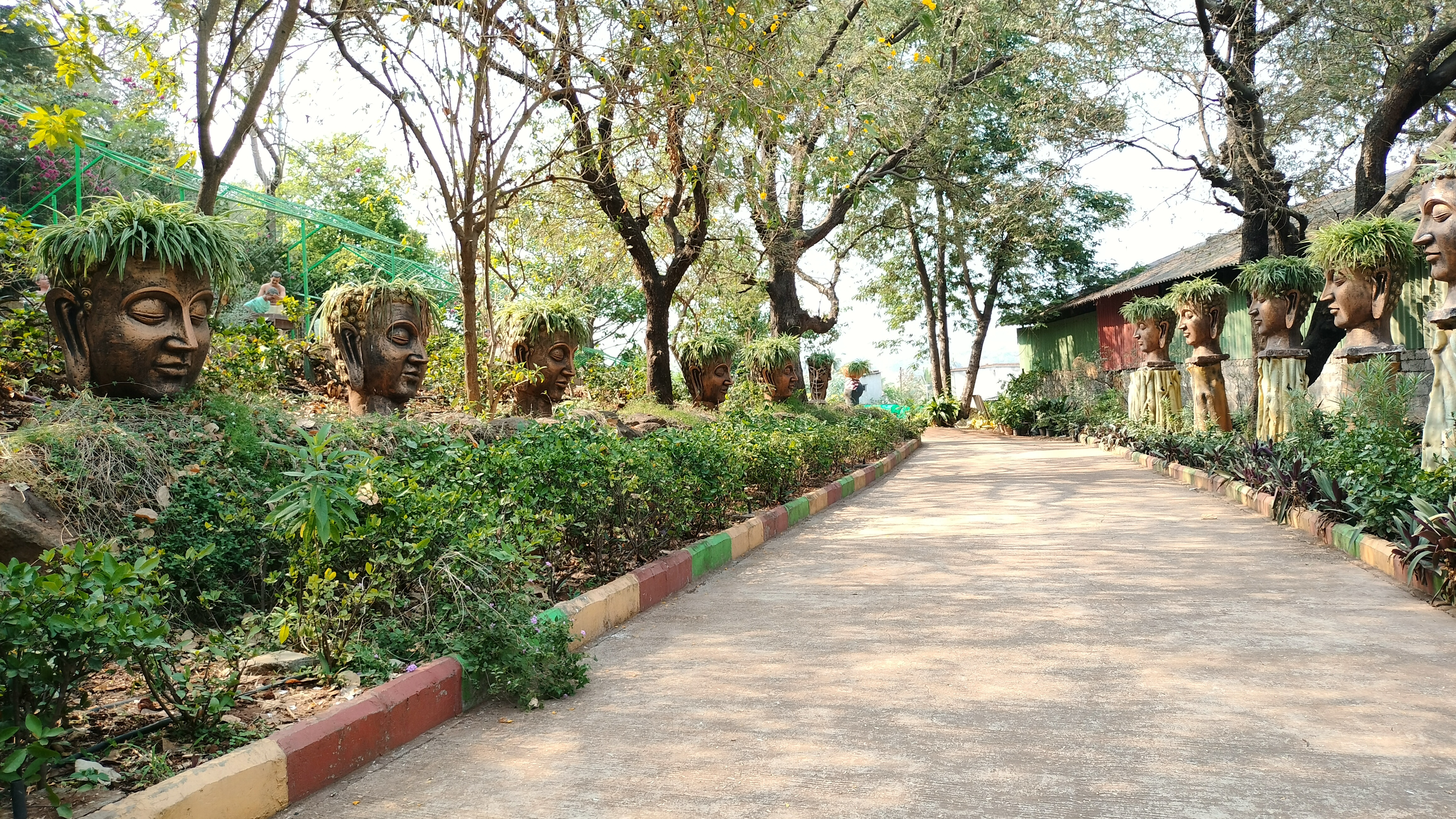 A path through a Divya garden with large artistic head sculptures on one side, each adorned with green plants as hair, and surrounded by trees and shrubbery.