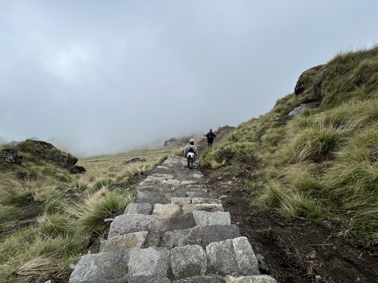 Two hikers descend a stone path through a grassy mountainous terrain, with the landscape shrouded in mist and fog near Khumai Dada, Kaski