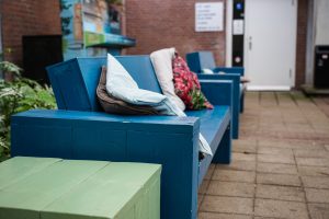 A close-up of blue wooden outdoor benches with various colorful cushions placed on them. The seating area is on a tiled patio adjacent to a brick wall. 