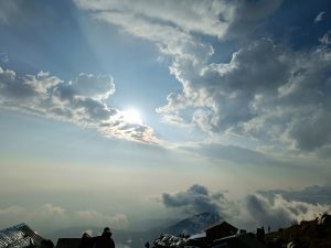 Scenic view of a mountain range with the sun partially obscured by clouds, casting rays of light across the sky. The foreground includes silhouetted rooftops and a few people observing the landscape. 