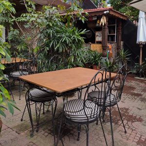 A cozy restaurant with a natural ambiance features a wooden dining table surrounded by ornate black metal chairs with striped cushions for four people, set in a tropical environment.