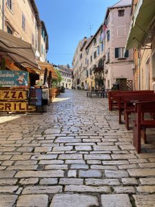  A quaint cobblestone street in a Croatian town is lined with old buildings on both sides. There are outdoor restaurant seating areas with wooden tables and chairs, and a sign for a pizza and grill restaurant. The buildings are adorned with shutters, and some have laundry hanging outside. The sky is clear and blue.