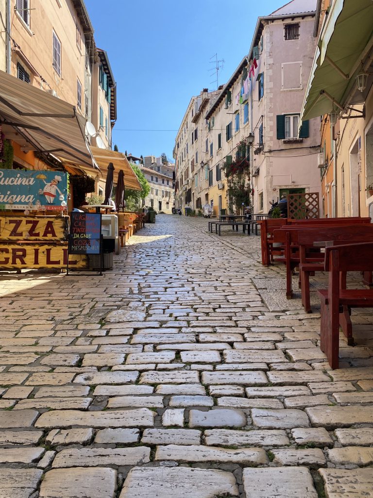 A quaint cobblestone street in a Croatian town is lined with old buildings on both sides. There are outdoor restaurant seating areas with wooden tables and chairs, and a sign for a pizza and grill restaurant. The buildings are adorned with shutters, and some have laundry hanging outside. The sky is clear and blue.