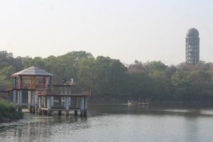 A lakeside scene featuring a pavilion on stilts extending over the water with people standing on it at Botanical Garden Lake at Dhaka, Bangladesh.