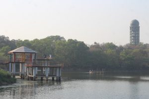 A lakeside scene featuring a pavilion on stilts extending over the water with people standing on it at Botanical Garden Lake at Dhaka, Bangladesh.
