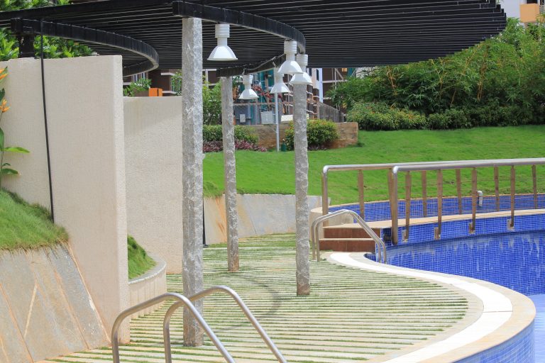 Outdoor scene with a swimming pool featuring a curved edge and stainless steel handrails, surrounded by a patterned tiled area and landscaped garden, complemented by a decorative light post and a sheltering structure.