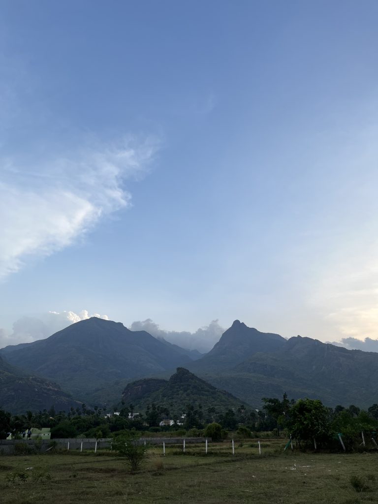 A serene landscape with a wide-open field in the foreground leading to a small fence, with a backdrop of majestic mountains under a vast sky with a few wispy clouds. Couralam, Tamilnadu.