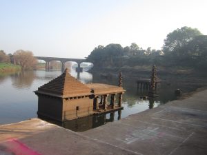 An ancient stone temple partially submerged in Panchganga river, with another smaller structure nearby. In the background, there is a bridge crossing the river and trees surrounding the area, creating a serene and historical atmosphere.