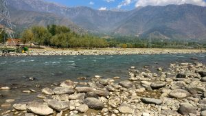 Clear, flowing river with large, smooth stones on its banks and bed. On the far side of the river is a grove of green trees, open fields, and a few scattered structures including a tractor with a green and yellow cart. In the background, towering mountains with rugged peaks rise under a partly cloudy blue sky.