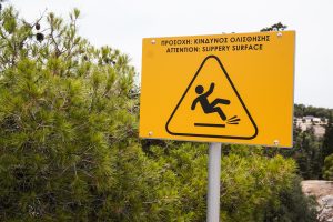 A yellow warning sign showing an icon of a person slipping, with the text "ATTENTION: SLIPPERY SURFACE" in English and a translation in Greek, placed next to green pine trees with a blurred background of a natural landscape.