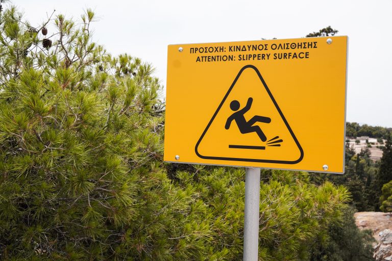 A yellow warning sign showing an icon of a person slipping, with the text “ATTENTION: SLIPPERY SURFACE” in English and a translation in Greek, placed next to green pine trees with a blurred background of a natural landscape.