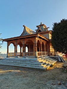 Temple under construction. A wooden building with intricate carvings and a steep roof, featuring a spacious porch and wide stone steps leading up to it, set against a clear blue sky. Trees flank the building on both sides.