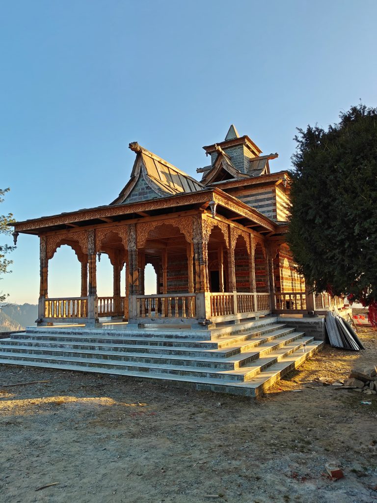 Temple under construction. A wooden building with intricate carvings and a steep roof, featuring a spacious porch and wide stone steps leading up to it, set against a clear blue sky. Trees flank the building on both sides.