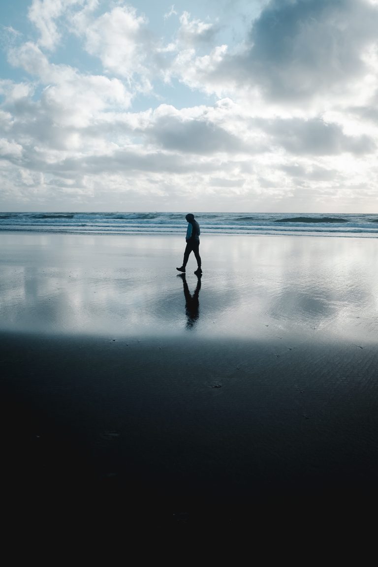 Person walking on the beach in the middle of a reflection of the clouds between the sky and the shore.