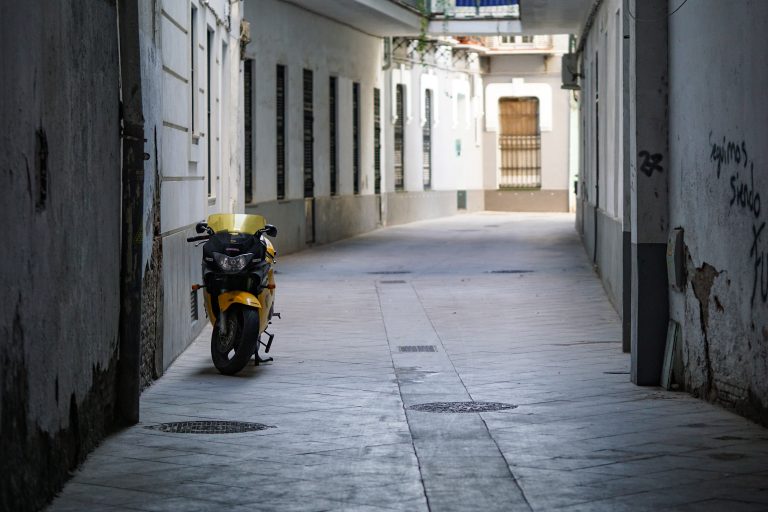 A yellow motorcycle parked on the sidewalk of a narrow urban alleyway with white buildings and graffiti on the walls.