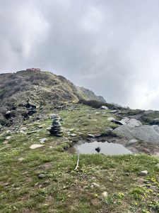 Kitchen Hill视点。草地小山上水坑旁的一小堆堆石头，背景是一座更大的小山。在较大的山上，在多云的天空下，可以看到山顶附近有一座红色的小建筑。