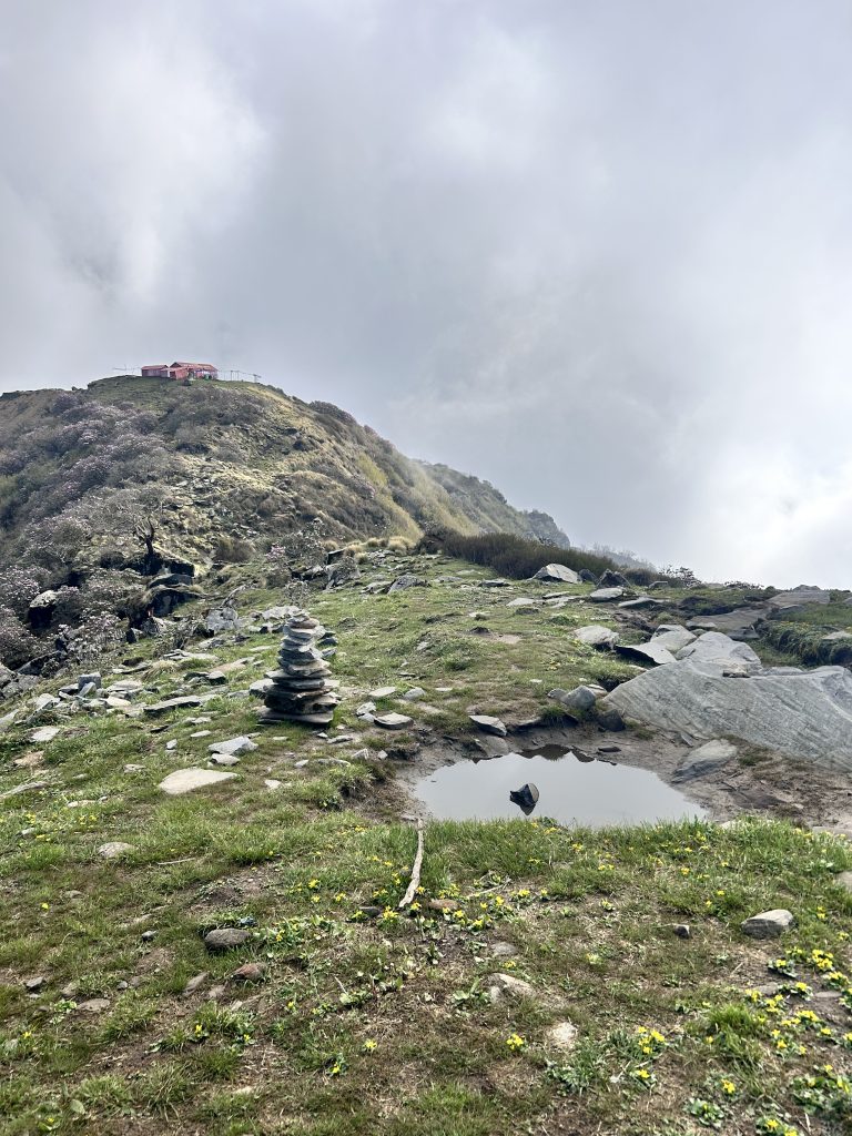 Kitchen Hill view point. A small pile of stacked rocks next to a puddle on a grassy hill, with a larger hill in the background. On the larger hill, a small red building is visible near the top under a cloudy sky.