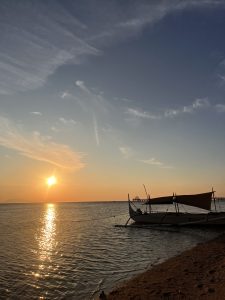 A serene sunset over a calm sea with vibrant orange light reflecting on the water. A small wooden boat is docked along the shoreline, with a pier and a gazebo visible in the distance along the horizon. Calatagan, Batangas, Philippines