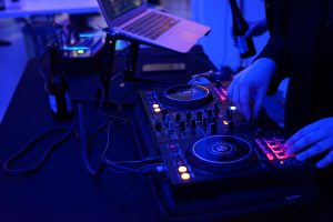 DJ's hands mixing a live session with digital plates and a laptop with a beer bottle on the table with purplish blue lighting.