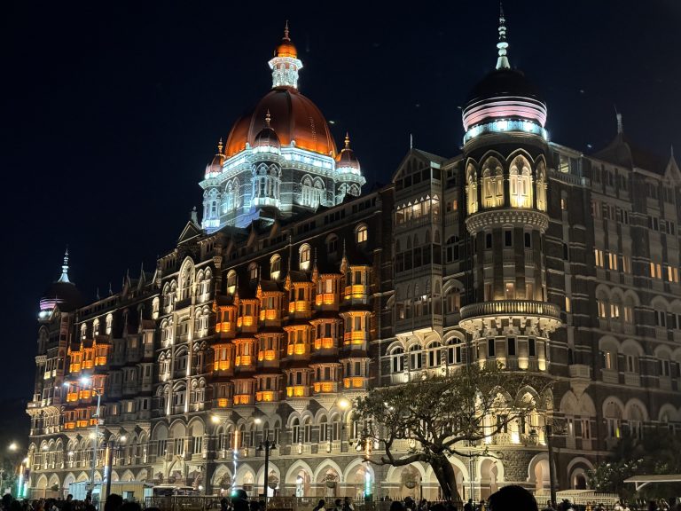 A nighttime view of Taj Mahal Palace hotel in Mumbai, an ornately designed, multi-story building with illuminated domes and arched windows. The building is lit up with warm orange and white lights, highlighting its intricate architectural details.