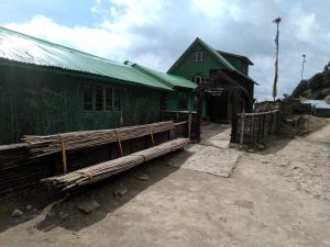 A green metal building on a rocky dirt road with two piles of bamboo poles on the side and a flagpole with blue, white, orange, and green flags on the other. A sign above the entrance says, “Habre’s Nest, Kaiyakata, Nepal, Tinchule Forest”
