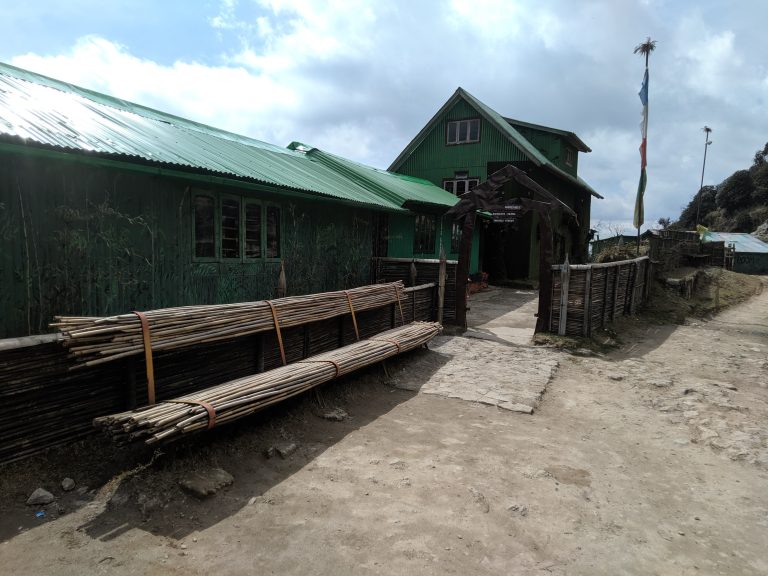 A green metal building on a rocky dirt road with two piles of bamboo poles on the side and a flagpole with blue, white, orange, and green flags on the other. A sign above the entrance says, "Habre's Nest, Kaiyakata, Nepal, Tinchule Forest"
