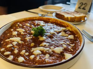 A close-up of a bowl of lasagna soup topped with a dollop of cheese and garnished with parsley. In the background, there is a plate with toast and a table number.