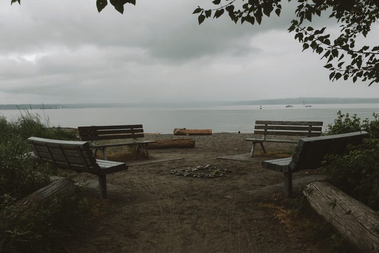 Set of four benches in a circle on a beach with views of the ocean in the background, along with several boats.