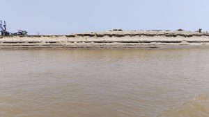 A view across a wide river with muddy water leading to a sandy cliff bank under a clear blue sky. Some vegetation and machinery are barely visible atop the distant bank.
