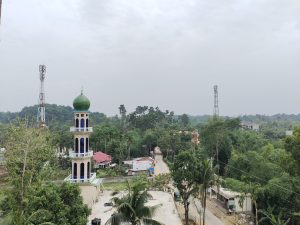 Tall minaret of a mosque featuring a green dome and multiple arched windows in the foreground. The background includes a road, scattered buildings, and trees, along with two communication towers.