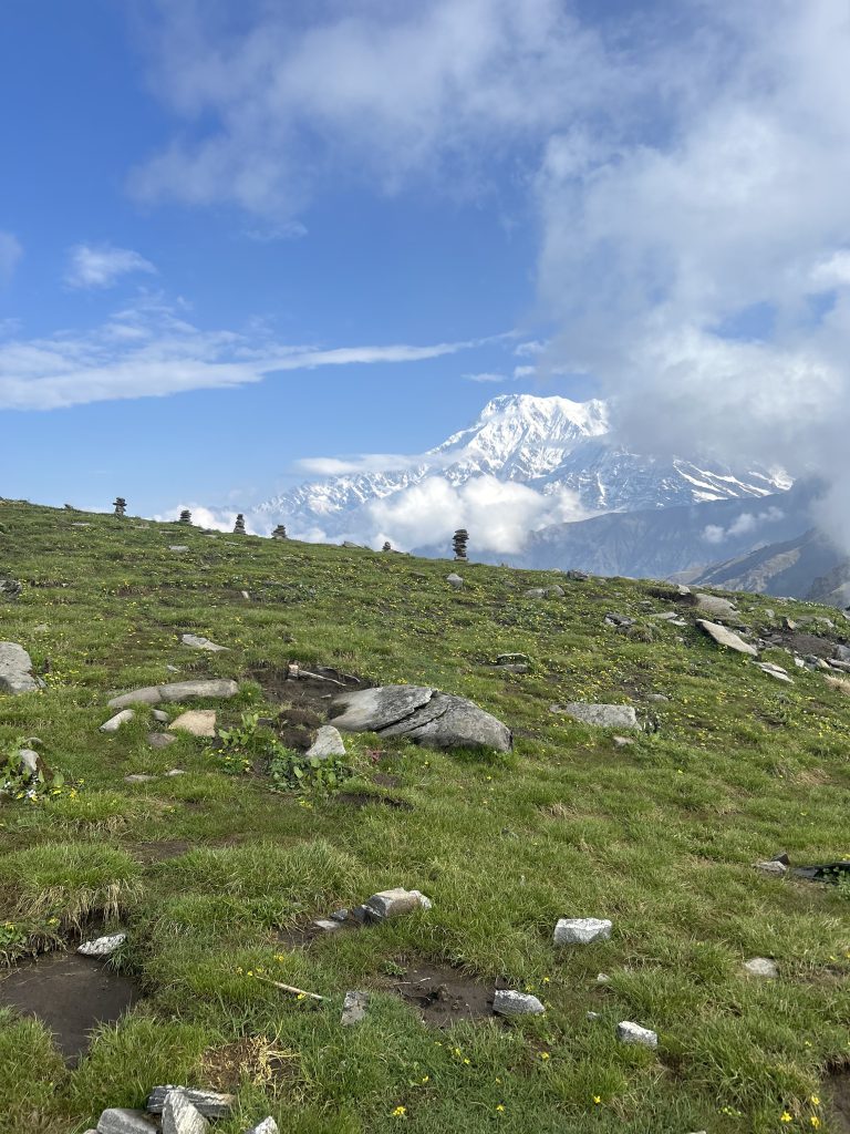 A lush green hillside dotted with rocks and small patches of flowers in the foreground, with a snow-capped mountain partially shrouded in clouds in the background under a bright blue sky.