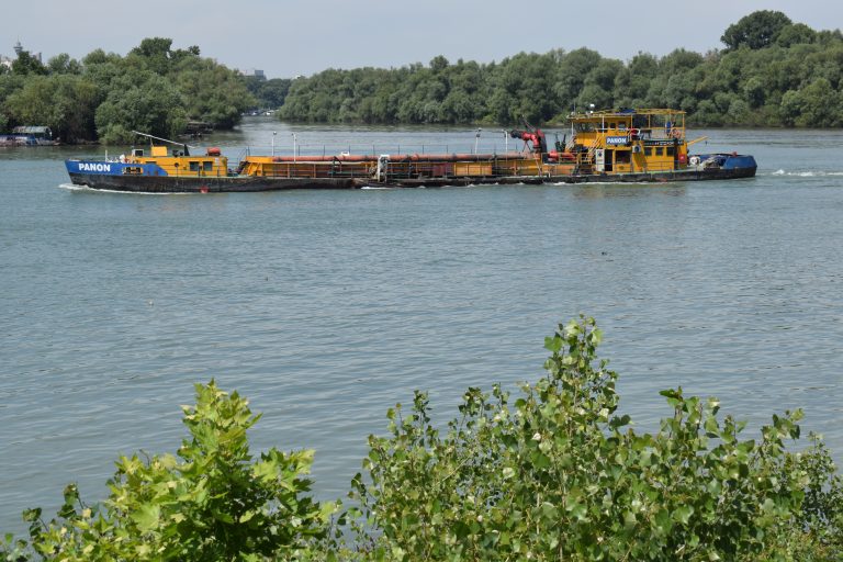 A yellow and blue barge traveling on a river with trees and foliage in the foreground and background.