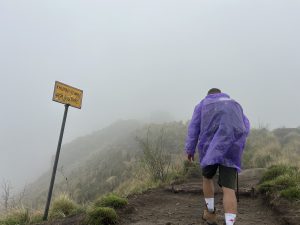 A person wearing a purple rain poncho is hiking up a misty trail. A yellow sign on the left indicates the destination "Khumai (45 mins)". The trail is surrounded by grassy terrain and low visibility due to the fog.