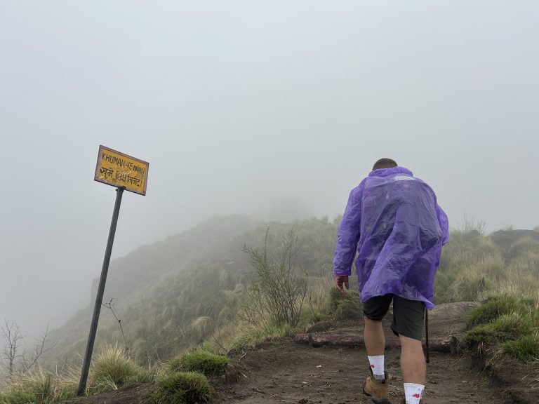 A person wearing a purple rain poncho is hiking up a misty trail. A yellow sign on the left indicates the destination “Khumai (45 mins)”. The trail is surrounded by grassy terrain and low visibility due to the fog.