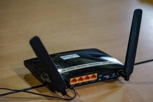 View larger photo: A black wireless router with two antennas on a wooden surface with visible status lights and a cable plugged in.