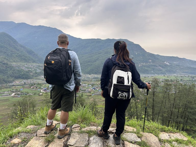 Two hikers with backpacks stand on a rocky outcrop overlooking a lush, green valley with scattered houses and surrounded by mountains under a cloudy sky.