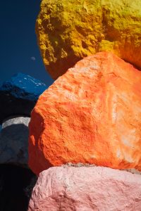 A stack of large, colorful rocks ranging from pink to blue to yellow with a view of the moon peeking through in a gap between them.