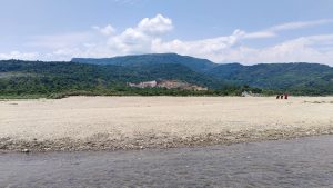 A wide view of a dry, rocky riverbed in the foreground with a small stream of water flowing through. In the background, there are lush green hills set against a partly cloudy sky. Four people, dressed in colorful clothing, are walking on the far right side of the riverbed.