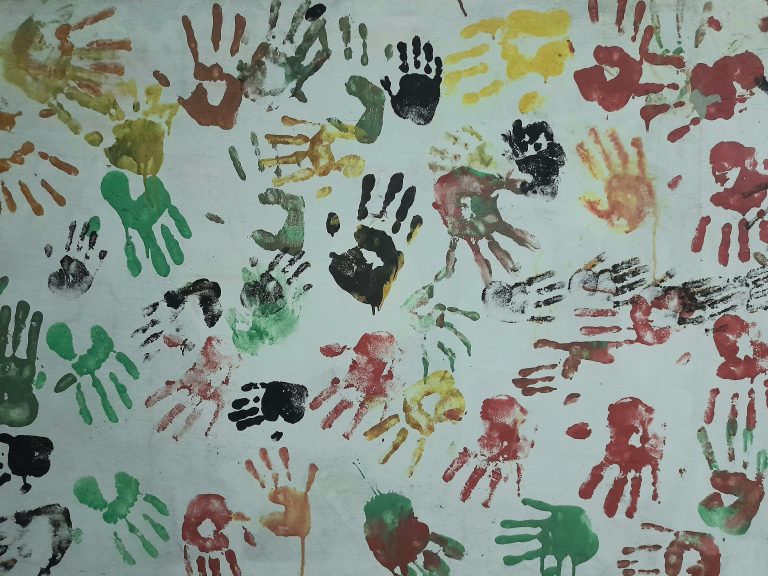 A white wall covered with colorful handprints in various shades of red, yellow, green, and black. The handprints are randomly placed and overlap in some areas.