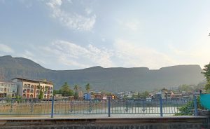 Town located near a body of water, with mountains in the background. The town has various buildings including colorful houses, palm trees, and a structure resembling a temple with flags on top. The sky is partly cloudy. In the foreground, there is a decorative metal railing along the edge of the water.
