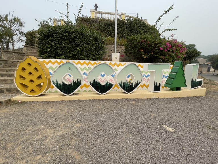 A colorful sign with large lettering on a pedestal, featuring graphic elements such as trees and architecture reflecting cultural identity, with lush greenery and flowering bushes in the background.
Wapu en Ocotal, NS